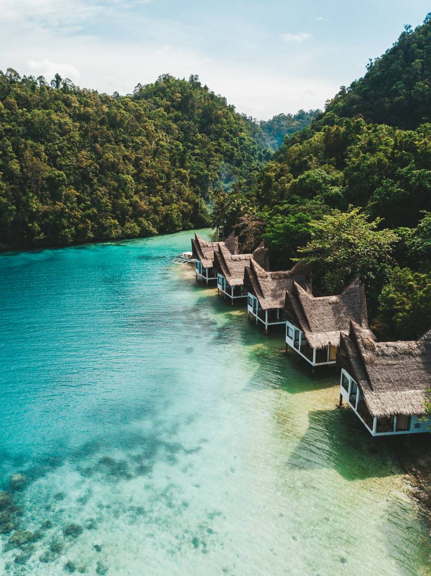 Thatched cottages on stilts along a turquoise inlet, backed by rainforest